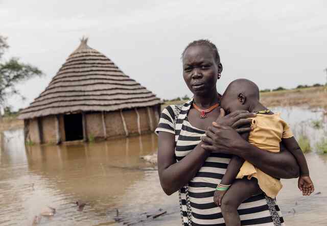 Abuk holds her child, Nyirou, outside of their flooded home. Water has flooded the area surrounding the home.